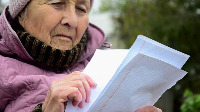 An old grandmother is reading a letter. The letter is in grandmother's old wrinkled hands. An old grandmother is reading close-up. An elderly woman reads outdoors. Letters in old hands