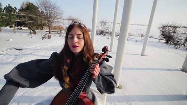 Young woman with diadem on her head plays cello with bow in winter. There is snow outside and the sun is shining