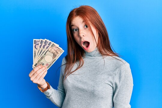 Young red head girl holding japanese yen banknotes scared and amazed with open mouth for surprise, disbelief face