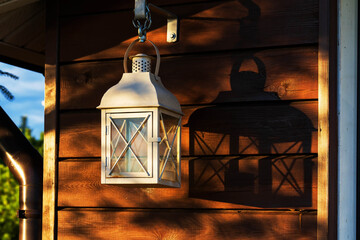 White metal lantern with a candle inside hangs against the wall of a brown wooden house. There is a shade from a lantern and a fragment of a downspout. Background.