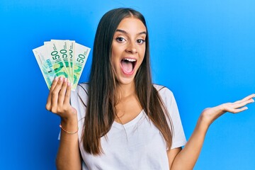 Young hispanic woman holding israel shekels celebrating achievement with happy smile and winner expression with raised hand