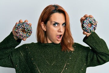 Hispanic young woman holding tasty colorful doughnuts in shock face, looking skeptical and sarcastic, surprised with open mouth