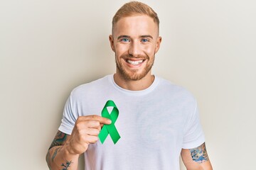 Young caucasian man holding support green ribbon looking positive and happy standing and smiling with a confident smile showing teeth