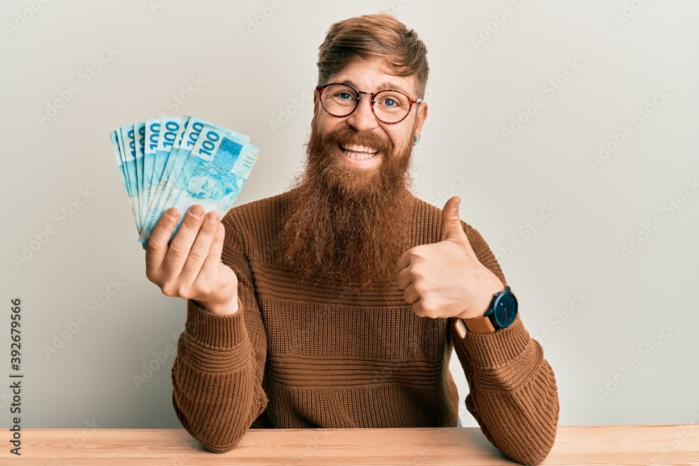 Canvas Prints Young irish redhead man holding 100 brazilian real banknotes sitting on the table smiling happy and positive, thumb up doing excellent and approval sign