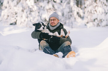 Fototapeta na wymiar Happy man holding lovely dog in his hands in snowy forest. Smiling boy hugging adorable puppy in winter wood. Pet lover. Dog - human`s friend concept.