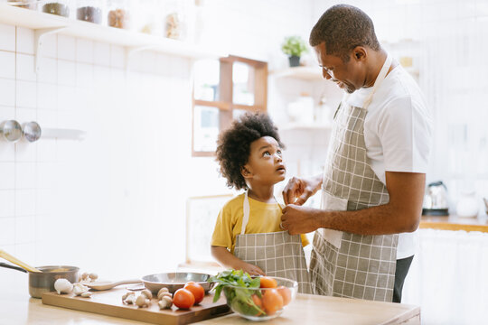 Happy African American father helping my son put on an apron before cooking in the kitchen at home. Spend weekend together, help, happy father and son concept.