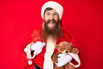Handsome young red head man with long beard wearing santa claus costume holding teddy bear smiling and laughing hard out loud because funny crazy joke.