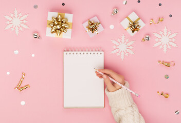 Woman hand holding pen and white empty notebook. Christmas festive pink background with gold gift boxes, decorations, snowflakes.