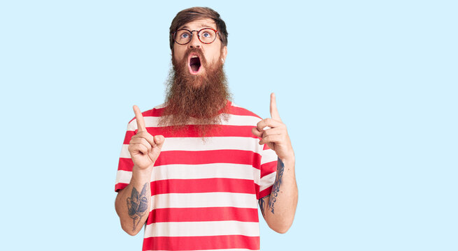Handsome young red head man with long beard wearing casual clothes amazed and surprised looking up and pointing with fingers and raised arms.