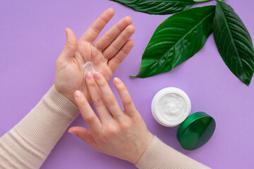 Young girls hand nearby white soft cream in jar with green leaves.Concept of eco cosmetic. Girl is moisturizing her hand by cream as beauty procedure at home.