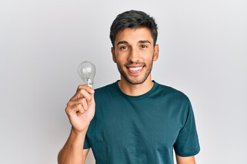Young handsome man holding lightbulb for inspiration and idea looking positive and happy standing and smiling with a confident smile showing teeth