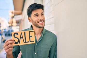 Young latin man smiling happy holding sale banner leaning on the wall at the city.