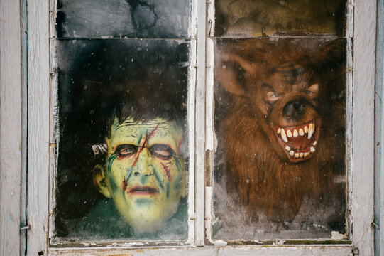 A child in a terrible monster mask peeks out of an old dirty window in winter