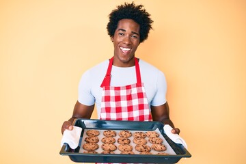 Handsome african american man with afro hair wearing baker uniform holding homemade cookies smiling...