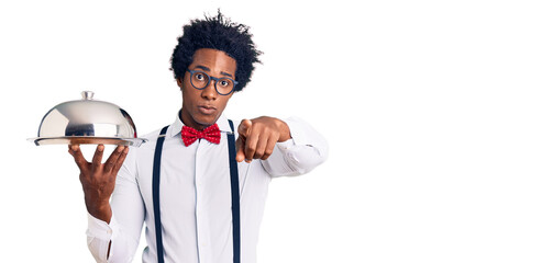 Handsome african american man with afro hair wearing waiter uniform holding silver tray pointing with finger to the camera and to you, confident gesture looking serious