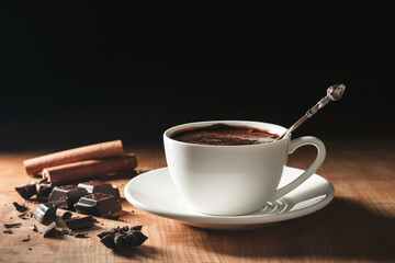 Hot chocolate drink in white cup, broken chocolate, cinnamon stick and star anise on a wooden table with dark background