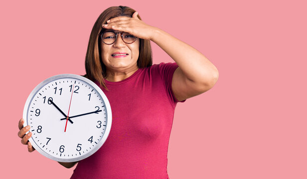 Middle age latin woman holding big clock stressed and frustrated with hand on head, surprised and angry face