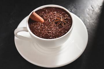 Hot chocolate drink in a white cup with cinnamon stick and star anise on textured dark background.