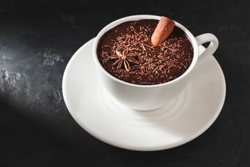 Hot chocolate drink in a white cup with cinnamon stick and star anise on textured dark background.