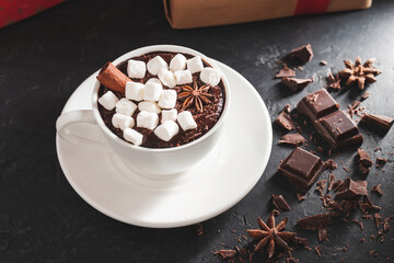 Hot chocolate drink in white cup with marshmallow, cinnamon stick, broken chocolate cubes and star anise on dark background