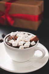 Hot chocolate drink in white cup with marshmallow and christmas gift box on dark background