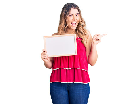 Young caucasian woman holding empty white chalkboard smiling happy pointing with hand and finger to the side