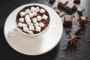 Hot chocolate drink in white cup with marshmallow, broken chocolate cubes and star anise on dark background