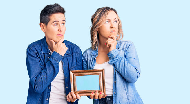 Couple of women holding empty frame thinking worried about a question, concerned and nervous with hand on chin