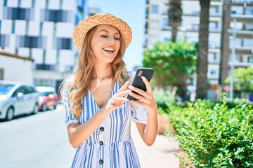 Young beautiful blonde woman on vacation wearing summer hat smiling happy. Standing with smile on face using smartphone at street of city.