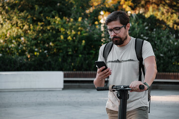 hipster man commuter with electric scooter outdoors in city, using smartphone.