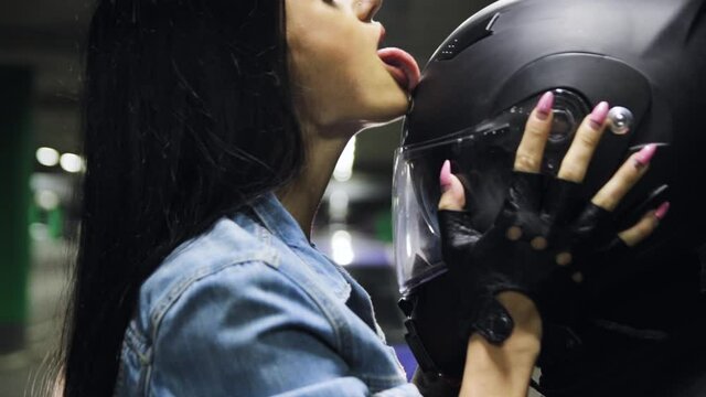 Young woman with long hair licks a black helmet of a motorcyclist in equipment with her tongue in an underground parking