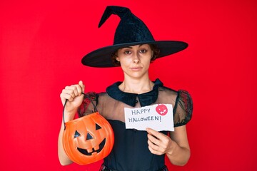 Young hispanic woman wearing witch costume holding pumpkin and happy halloween message skeptic and...