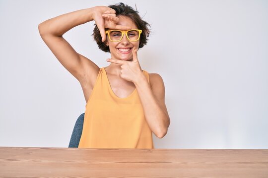 Young hispanic woman wearing casual clothes and glasses sitting on the table smiling making frame with hands and fingers with happy face. creativity and photography concept.