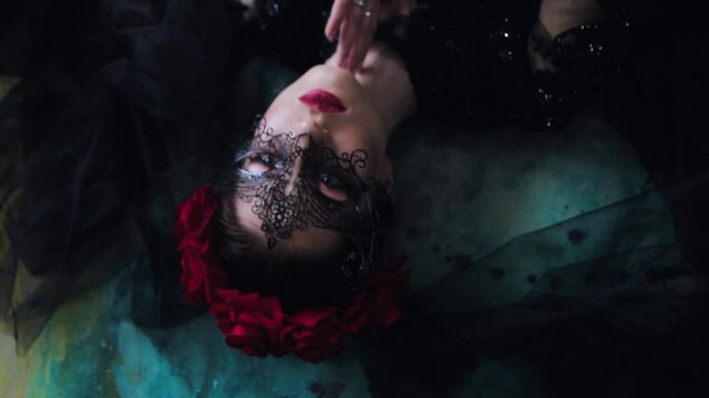 Beautiful young woman in a black masquerade mask and black peignoir lies on multi-colored floor
