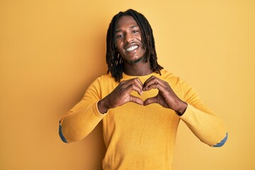 African american man with braids wearing casual yellow sweater smiling in love doing heart symbol shape with hands. romantic concept.