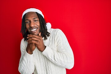African american man with braids wearing christmas hat laughing nervous and excited with hands on...