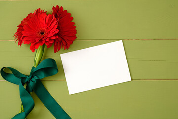 Bouquet of red gerberas and blank paper card on pastel green wooden background. Flat lay composition, greeting card mockup with flowers. Flat lay, top view, overhead.