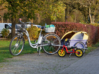 Fototapeta premium Silver Electric bicycle with a children's trailer. Parked in the park E-bike and child trailer. Small children's bike. A bicycle with a basket on the handlebars