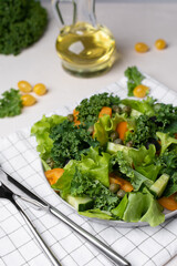 veggie salad of lettuce leaves and kale. Yellow tomatoes, oruganets, olives and capers. A plate on a checkered cloth napkin. A bottle of olive oil is nearby. Light vegetable salad. Light background.