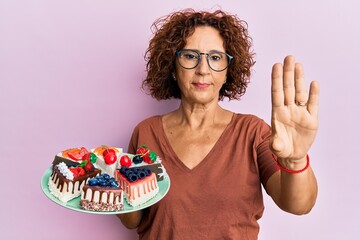 Beautiful middle age mature woman holding cake slices with open hand doing stop sign with serious...