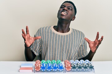 Young african american man sitting on the table with poker chips and cards crazy and mad shouting and yelling with aggressive expression and arms raised. frustration concept.