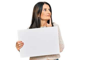 Middle age brunette woman holding blank empty banner serious face thinking about question with hand on chin, thoughtful about confusing idea