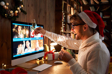 Young man wearing Christmas hat drinking champagne holding sparkler talking to friends on virtual...