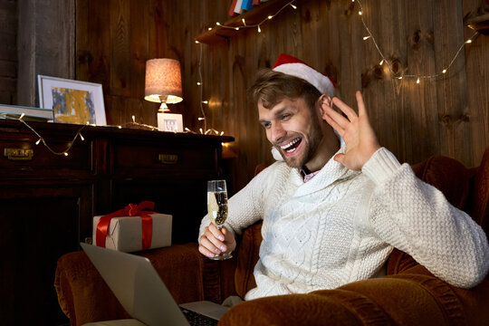 Happy young man wearing santa hat waving hand celebrating virtual New Year party using laptop holding champagne glass and Christmas gift having distance online chat meeting with friends at home.