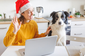 Dog and woman wearing Santa hat waving hand video calling family by webcam. Girl with laptop having virtual meeting chat on holidays sitting on kitchen at home Happy Christmas and New Year new normal