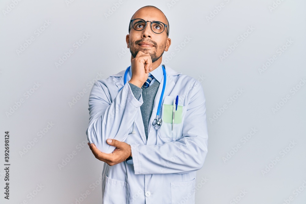 Sticker Hispanic adult man wearing doctor uniform and stethoscope with hand on chin thinking about question, pensive expression. smiling and thoughtful face. doubt concept.