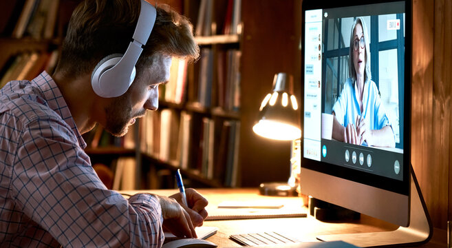 Male student wearing headphones conference video calling, watching webinar, online training class, virtual chat meeting with remote teacher or coach distance learning using computer, taking notes.