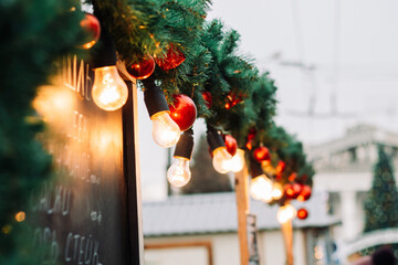 Christmas market's decorations closeup. Outdoor garland lights on a branch of fir tree with red...