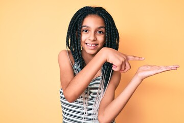 Young african american girl child with braids over yellow background amazed and smiling to the camera while presenting with hand and pointing with finger.