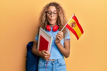 Beautiful caucasian teenager girl exchange student holding spanish flag making fish face with mouth...